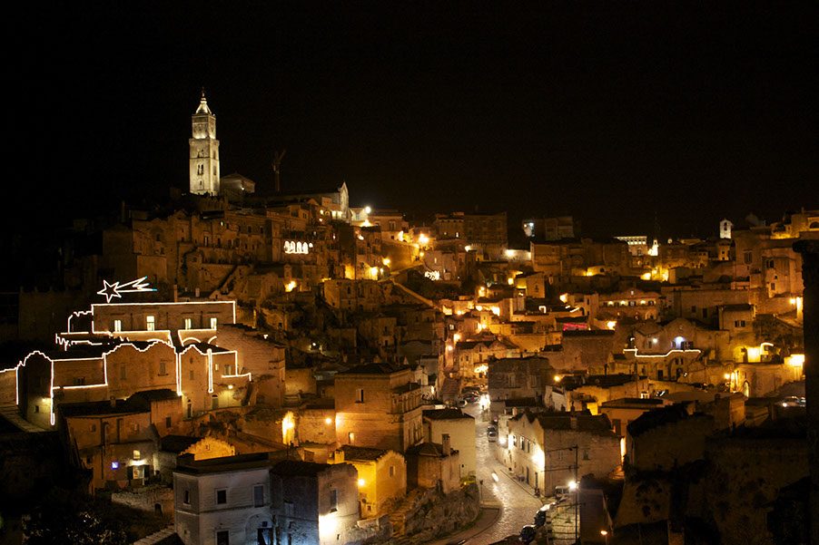 Matera at night, the lit town climbing the hillside under a black sky.
